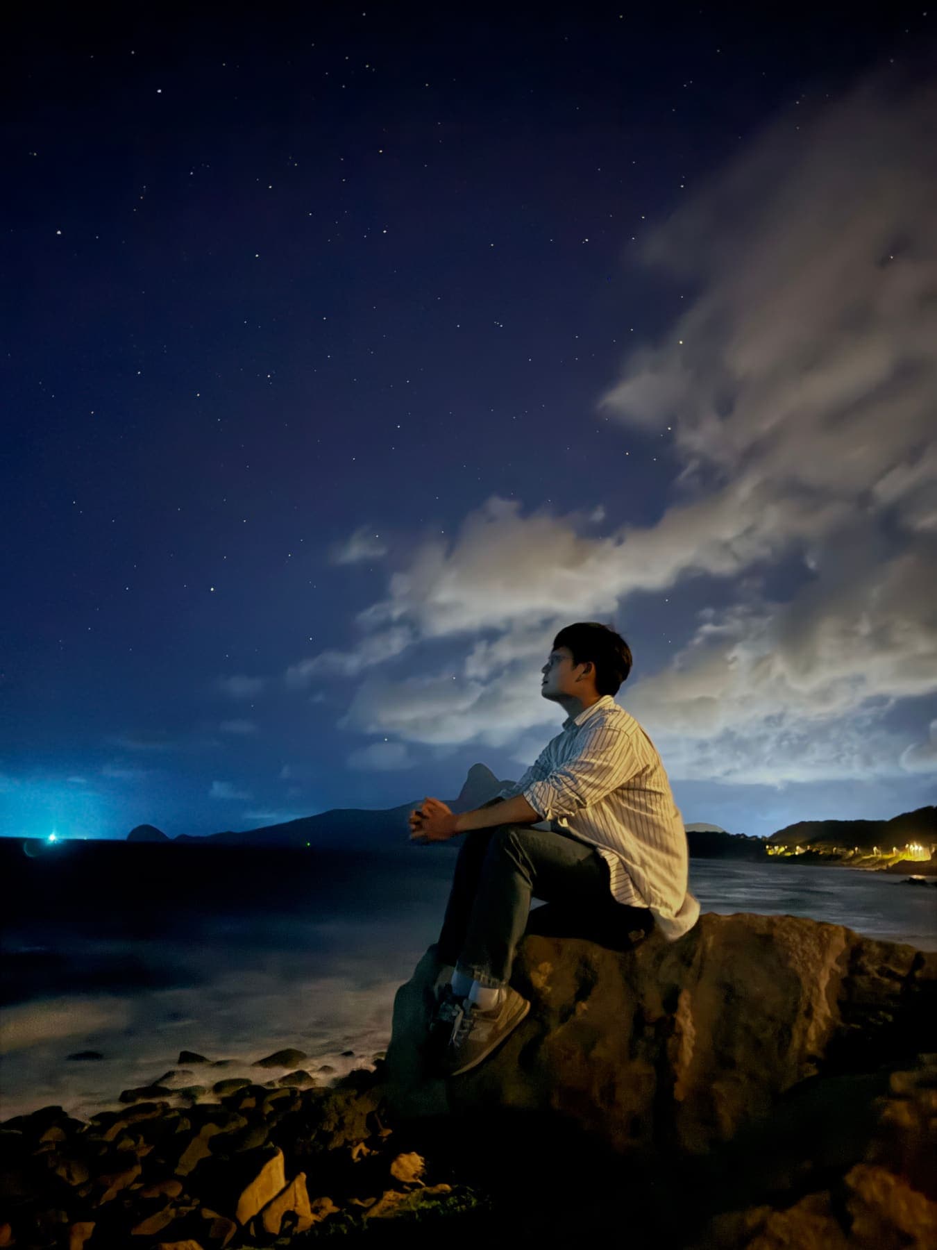 A man sitting on a seaside rock at night, looking up at the stars