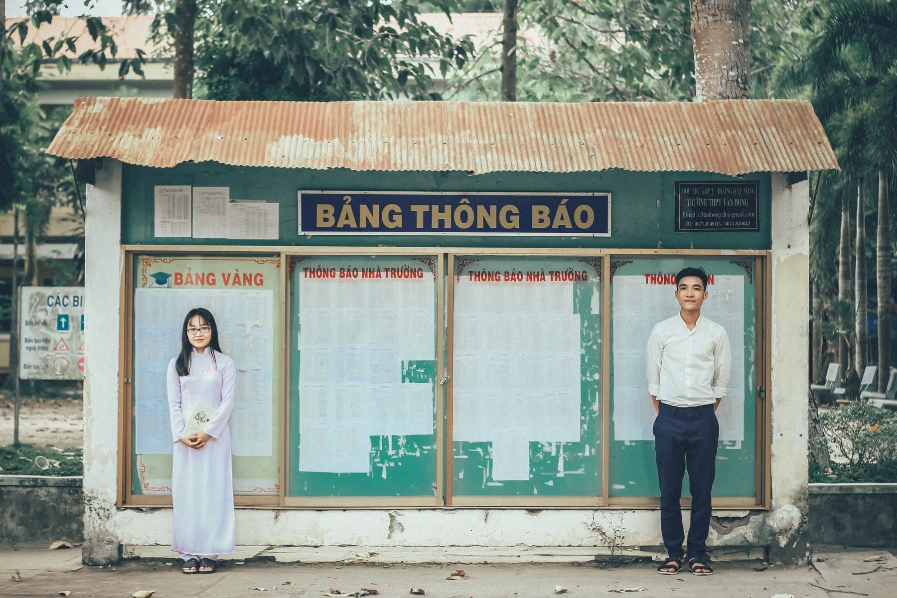 A girl in a white áo dài and a boy in a white shirt beside a school notice board