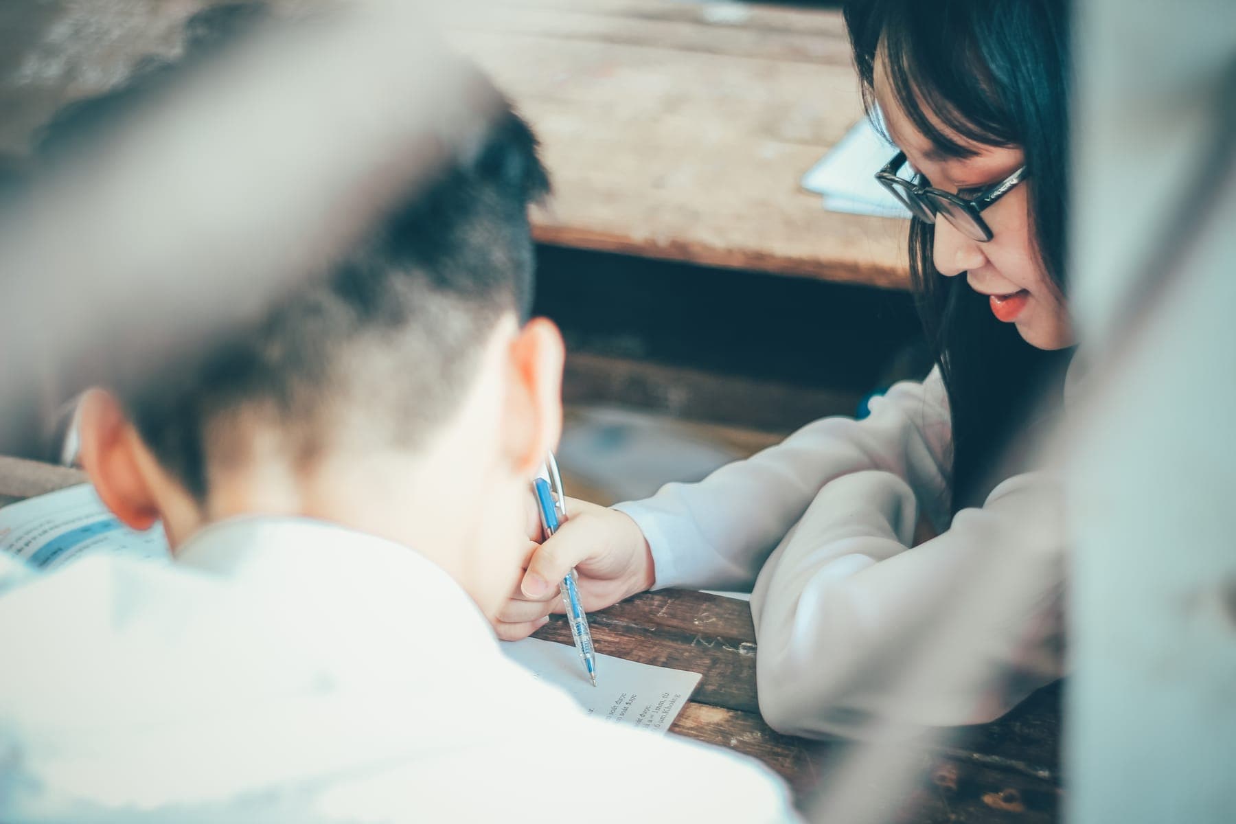 A girl in glasses writing beside a boy, framed by a blurred desk