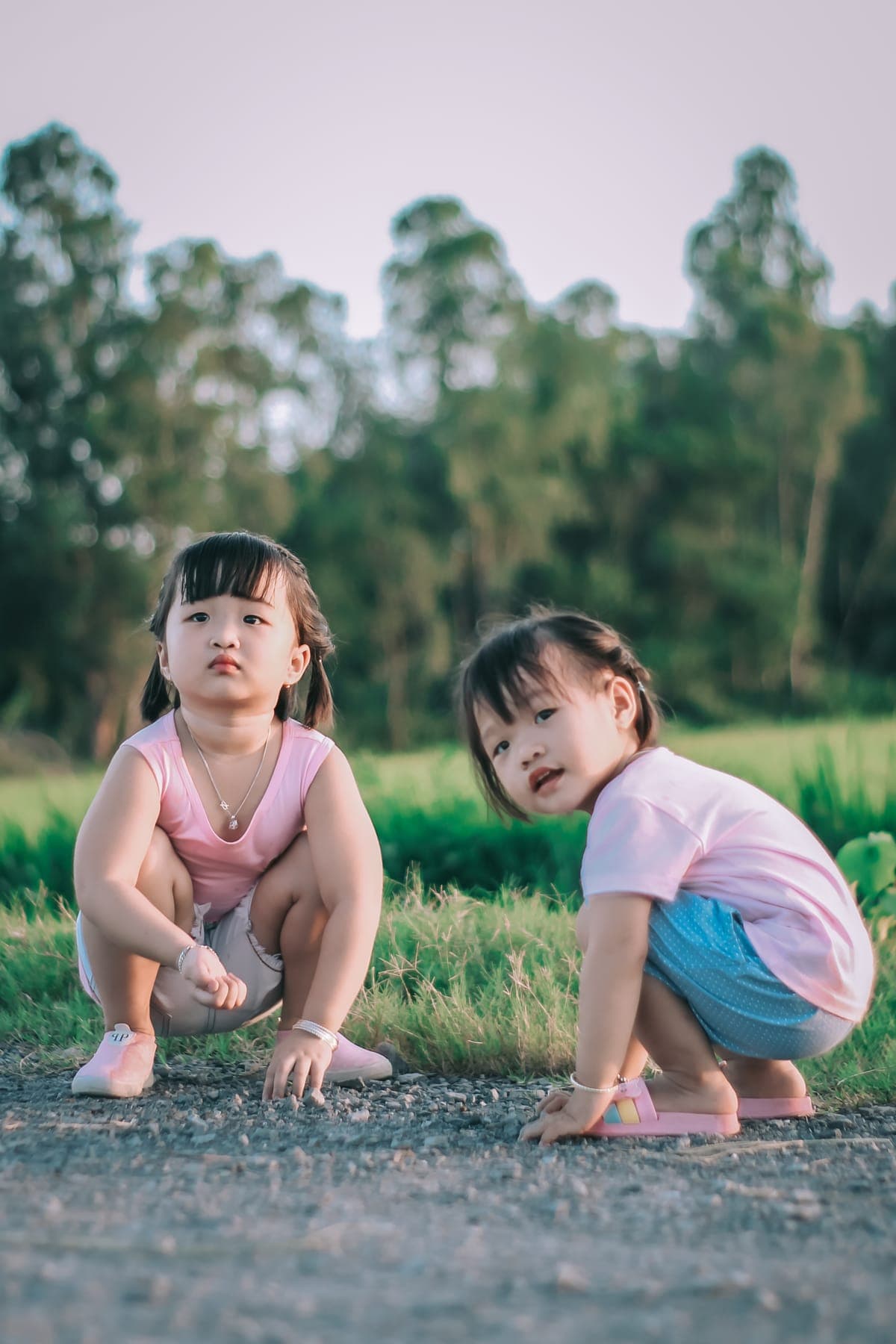 Two little girls crouching on a gravel path at the edge of a field